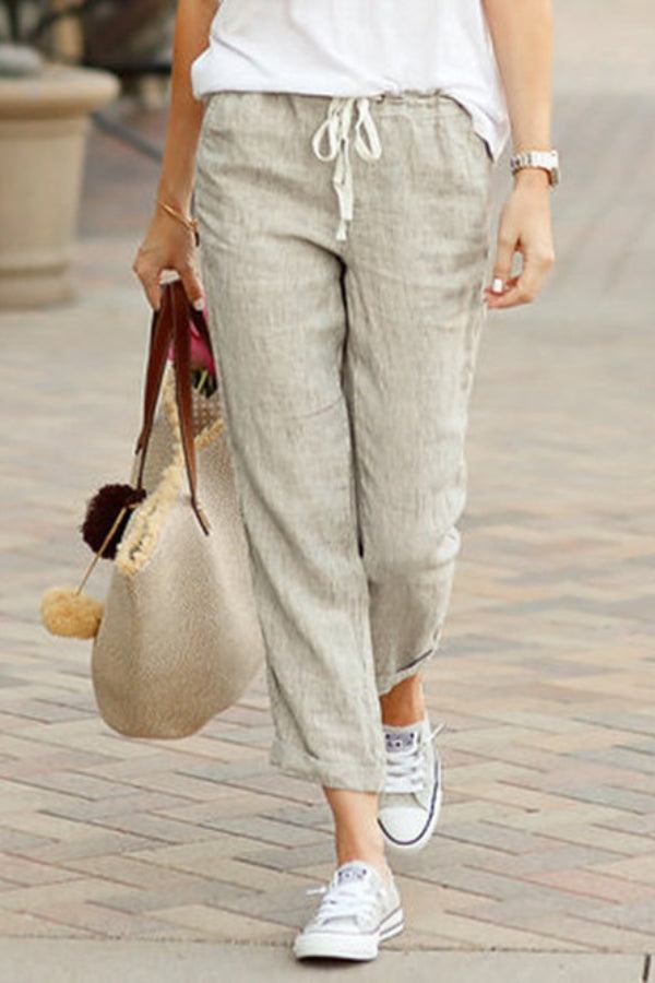 Woman wearing beige relaxed fit straight pants with drawstring waist and white sneakers walking outdoors carrying a woven tote bag