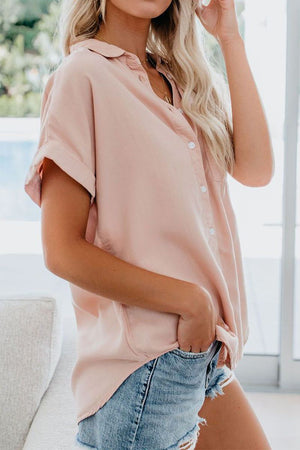 Woman wearing a soft pink V-neck collared button blouse with rolled short sleeves and denim shorts