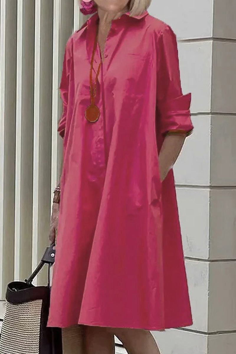 Woman wearing a vibrant pink long shirt-style dress with rolled sleeves and collar, accessorized with a long necklace and handbag outdoors.