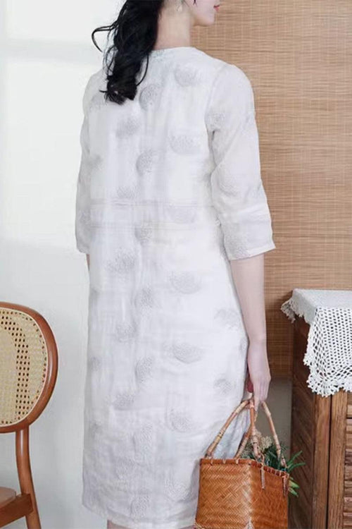 Back view of a woman wearing a white 100% linen embroidered notch-neck midi dress holding a woven bag indoors