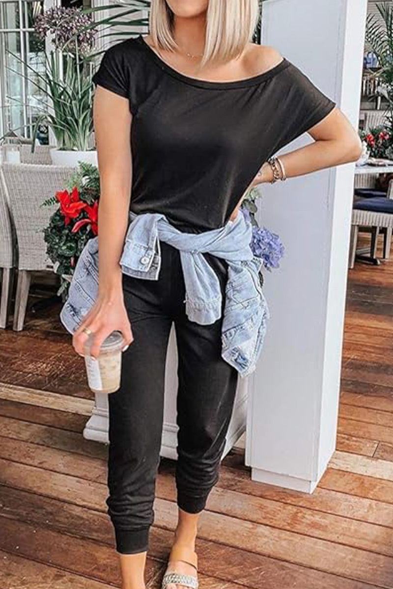 Woman wearing black Moonlit Rome Off-Shoulder jumpsuit with denim jacket tied around waist, standing indoors with drink in hand