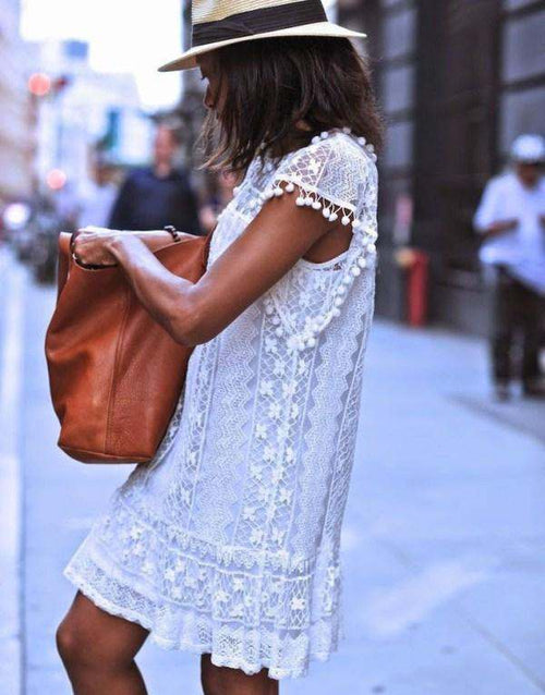 Woman wearing white pom pom lace mini dress with sleeveless design and carrying a brown bag on city street