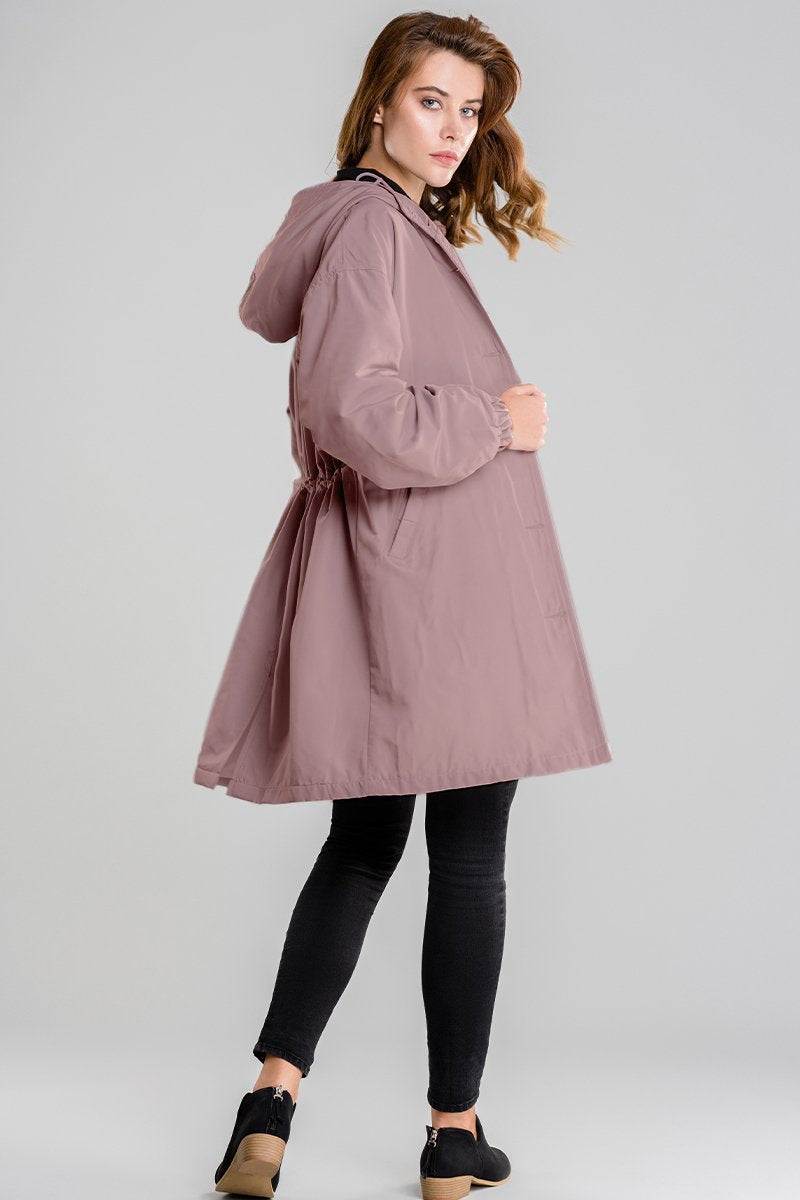 Woman wearing pink water resistant oversized hooded windbreaker jacket with adjustable waist and long length over black pants and black shoes.