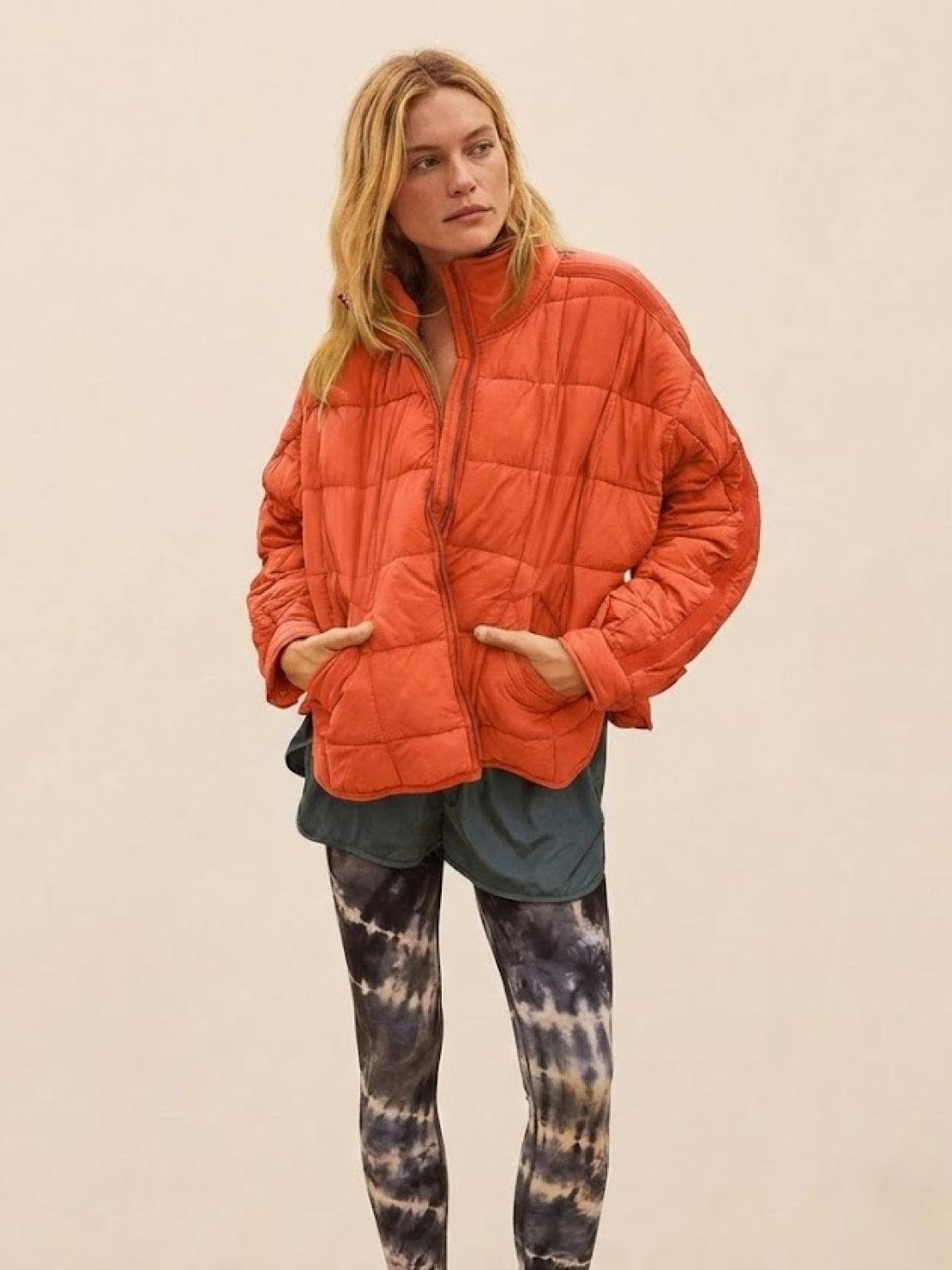Lightweight puffer travel jacket in orange with quilted design, worn by woman with hands in pockets.