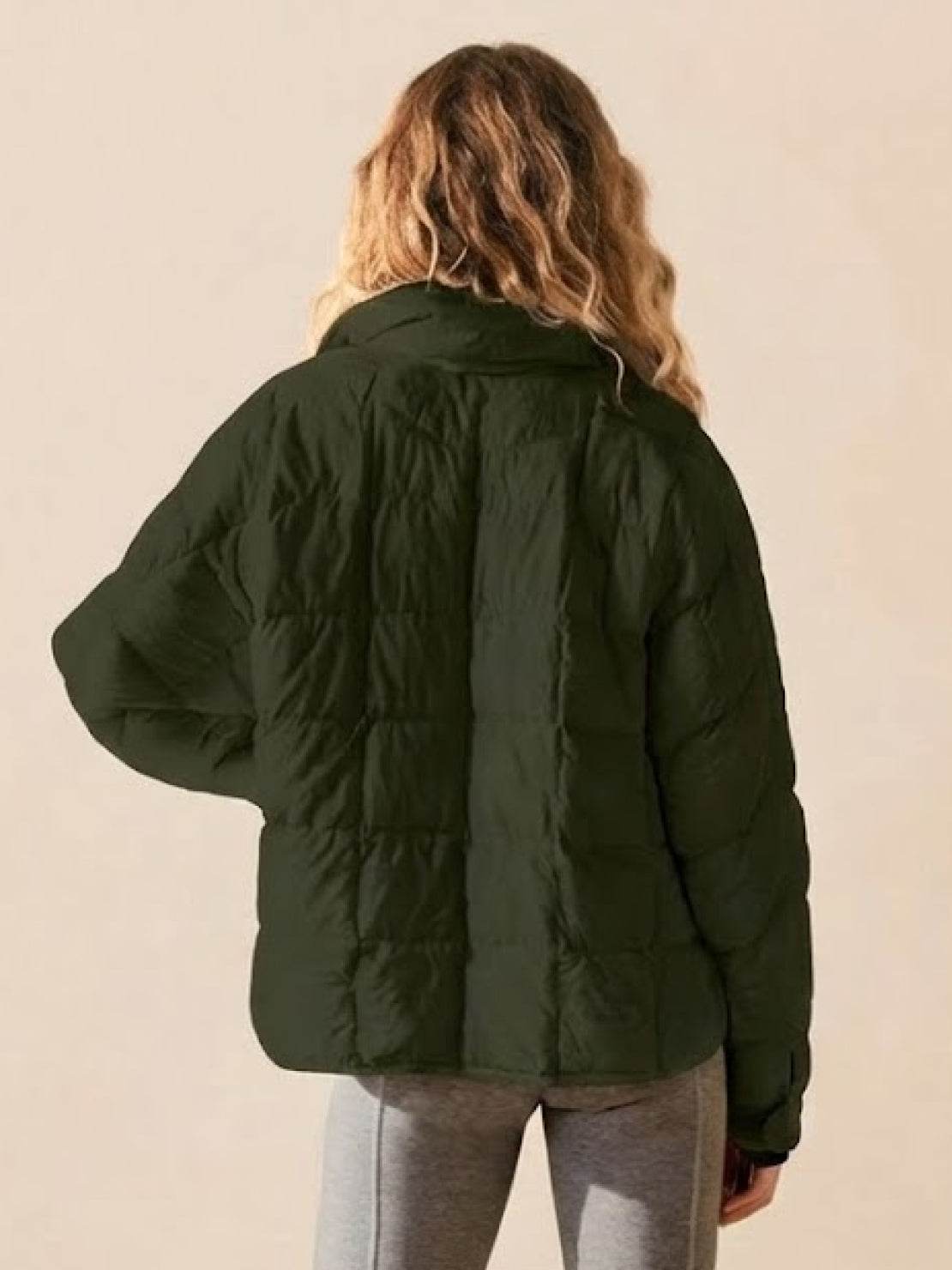 Back view of woman wearing dark green lightweight puffer travel jacket and gray pants against beige background