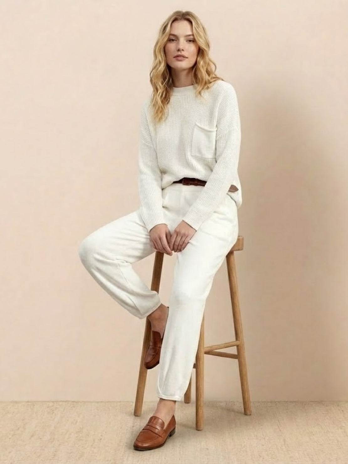 Woman wearing European minimalism relaxed knit sweater top and light drapey trousers sitting on wooden stool.