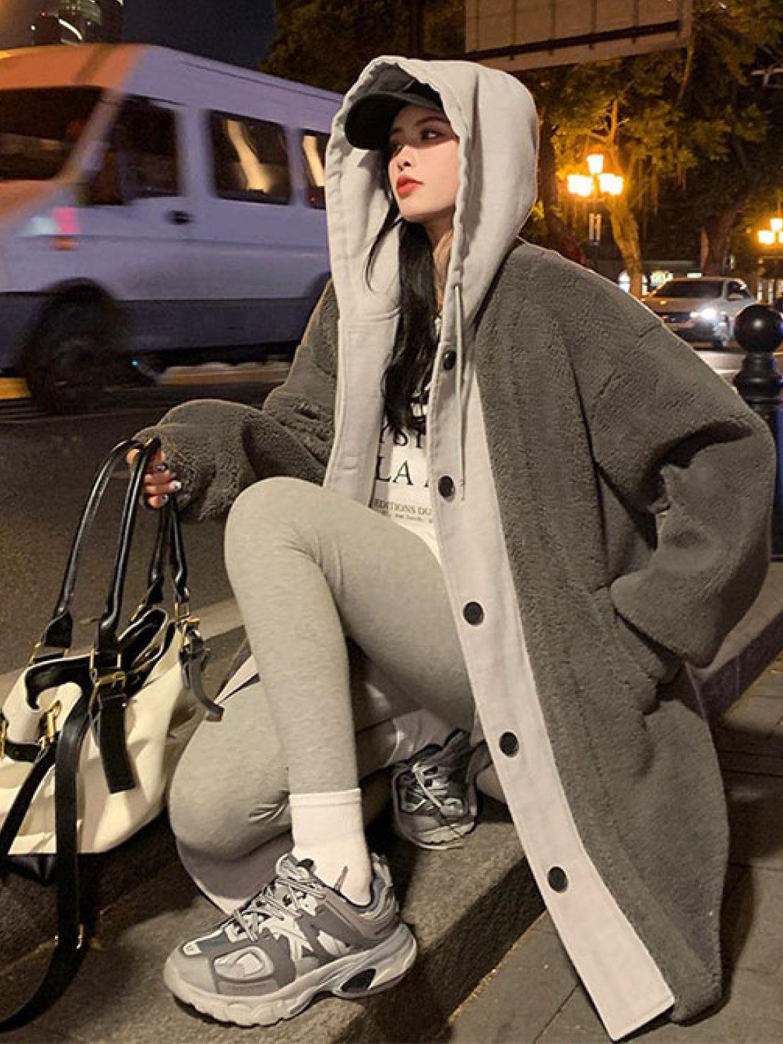 Woman wearing oversized gray sherpa coat with hood, paired with gray leggings and sneakers, holding a tote bag sitting outdoors at night