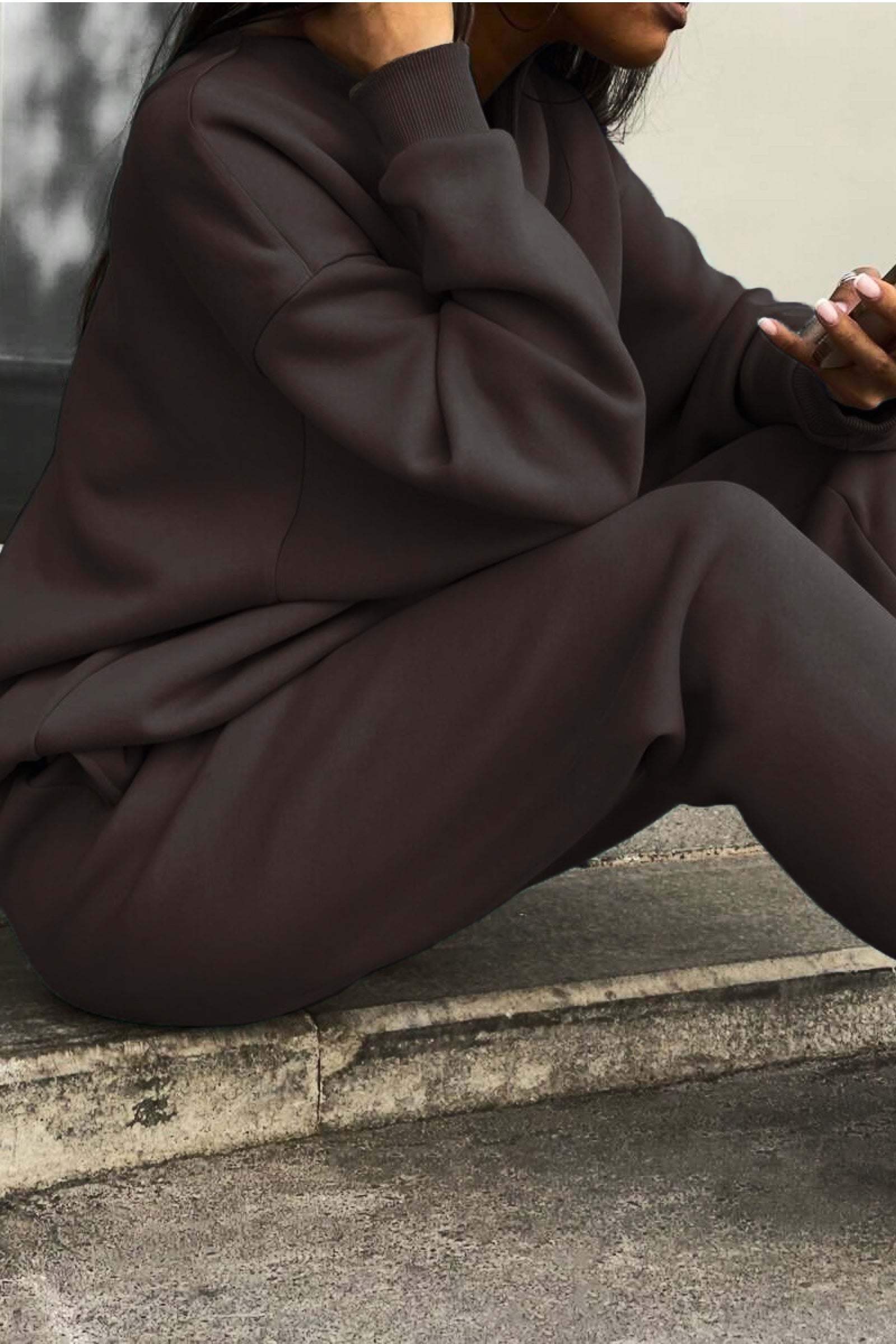 Woman wearing a dark brown cotton blend tracksuit sitting on concrete steps, showing relaxed fit and soft texture.