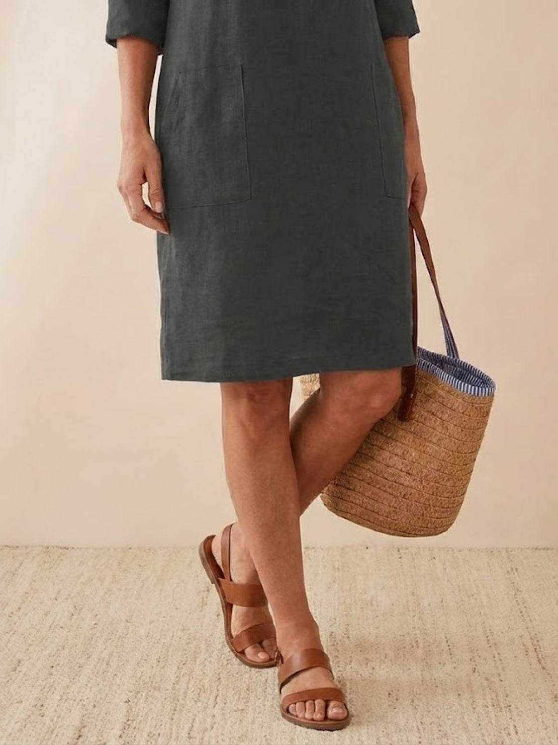 woman wearing knee-length dark cotton dress with rolled 3/4 sleeves and brown sandals holding woven basket bag