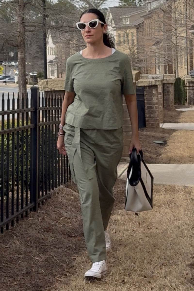 Woman wearing olive green cotton linen set with short sleeve top and straight-leg pants walking outdoors carrying tote bag