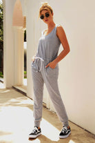 Woman wearing grey comfy sleeveless vest pocket jumpsuit with elastic waistband and drawstring, styled casually with sunglasses and black sneakers.