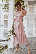 Woman wearing Elegant Blossoms Boho Maxi Dress with floral pattern, V-neck, belted waist, and ruffled hem holding flowers on a wooden porch.