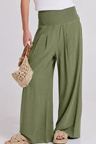 Woman wearing olive green palazzo pants with a smocked waistband holding a woven bag, paired with sandals.