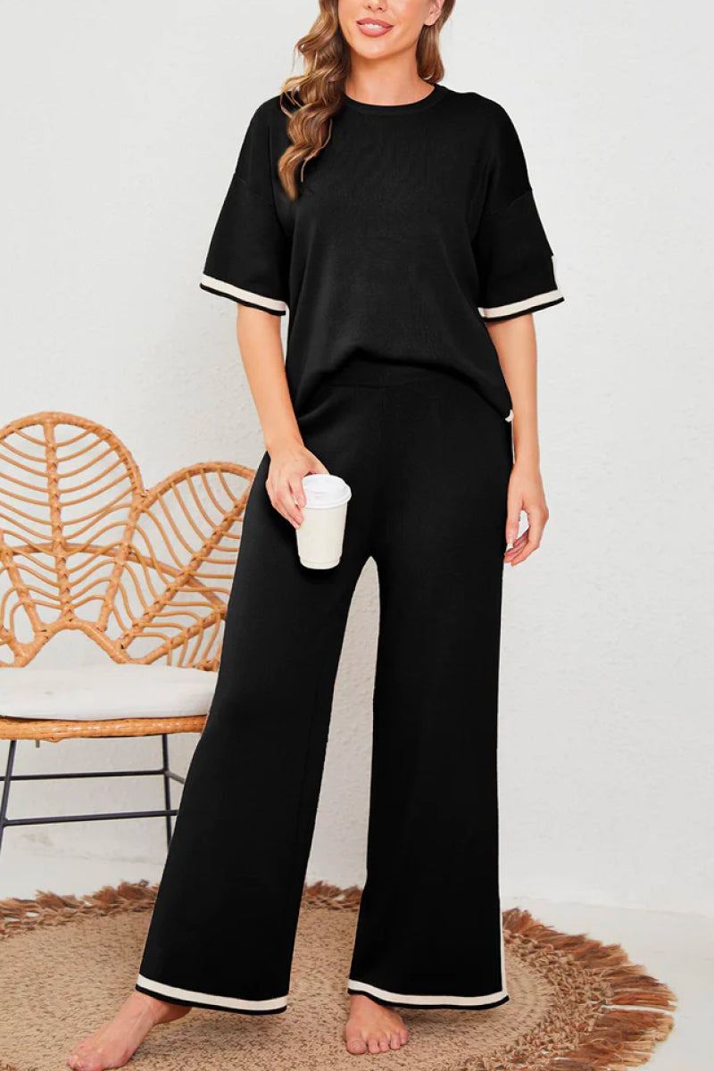 Woman wearing a black short sleeved relaxed fit lounge two-piece set with wide-leg pants and white trim on sleeves and hem, holding a cup.