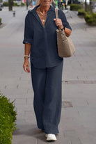 Woman wearing a navy relaxed shirt and trousers two-piece set with rolled-up sleeves and white sneakers, carrying a beige woven bag, walking outdoors.
