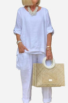 Woman wearing white asymmetrical two-piece trouser set with rolled sleeves and holding woven handbag