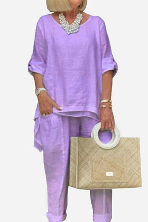 Woman wearing lavender asymmetrical two-piece trouser set with rolled sleeves holding woven handbag