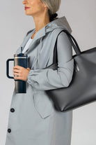 Woman holding indestructible travel cup with handle and secure lid, wearing gray jacket and carrying black tote bag