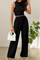 Woman wearing black Stroll & Hangout Ribbed Knit sleeveless top and wide-leg pants set holding white handbag, indoor plant background