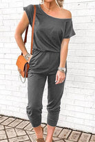 Woman wearing Moonlit Rome Off-Shoulder black jumpsuit with brown shoulder bag against white brick wall