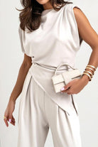 Woman wearing Manhattan Lines Draped sleeveless asymmetrical top and wide-leg pants set in beige with a small handbag and gold bracelets