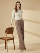 Woman wearing brown thermal wool blend trousers with a high waist and wide-leg silhouette, paired with a cream turtleneck sweater, standing next to a wooden chair in a minimalist setting.