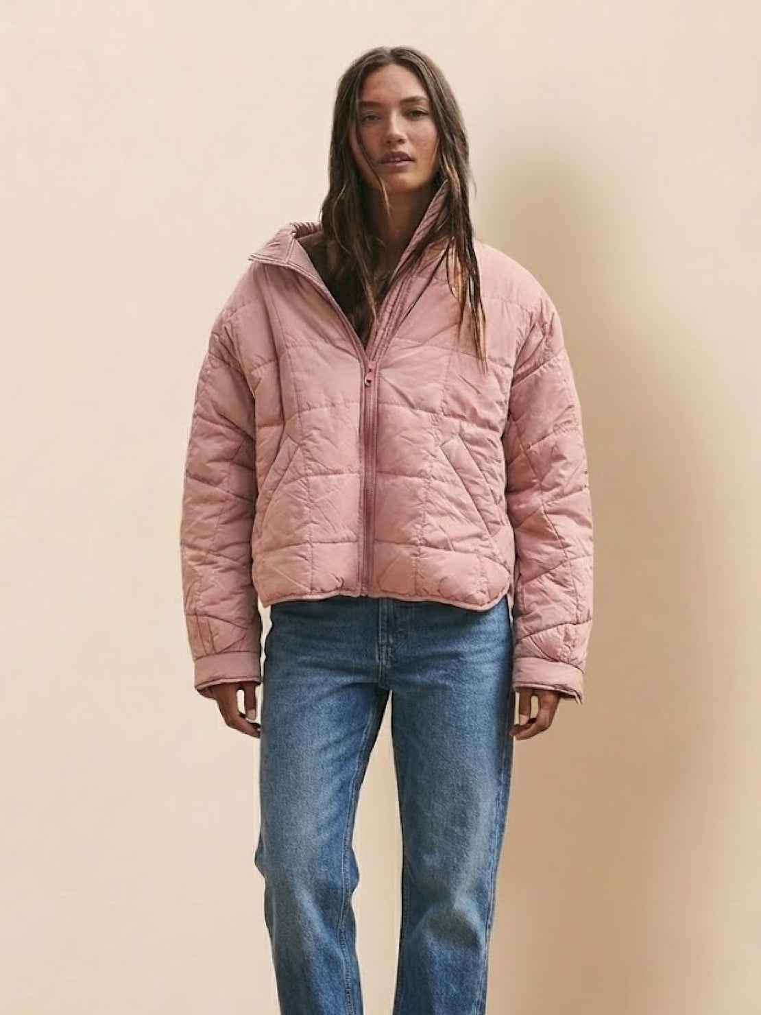Woman wearing a pink lightweight puffer travel jacket with jeans against a neutral background