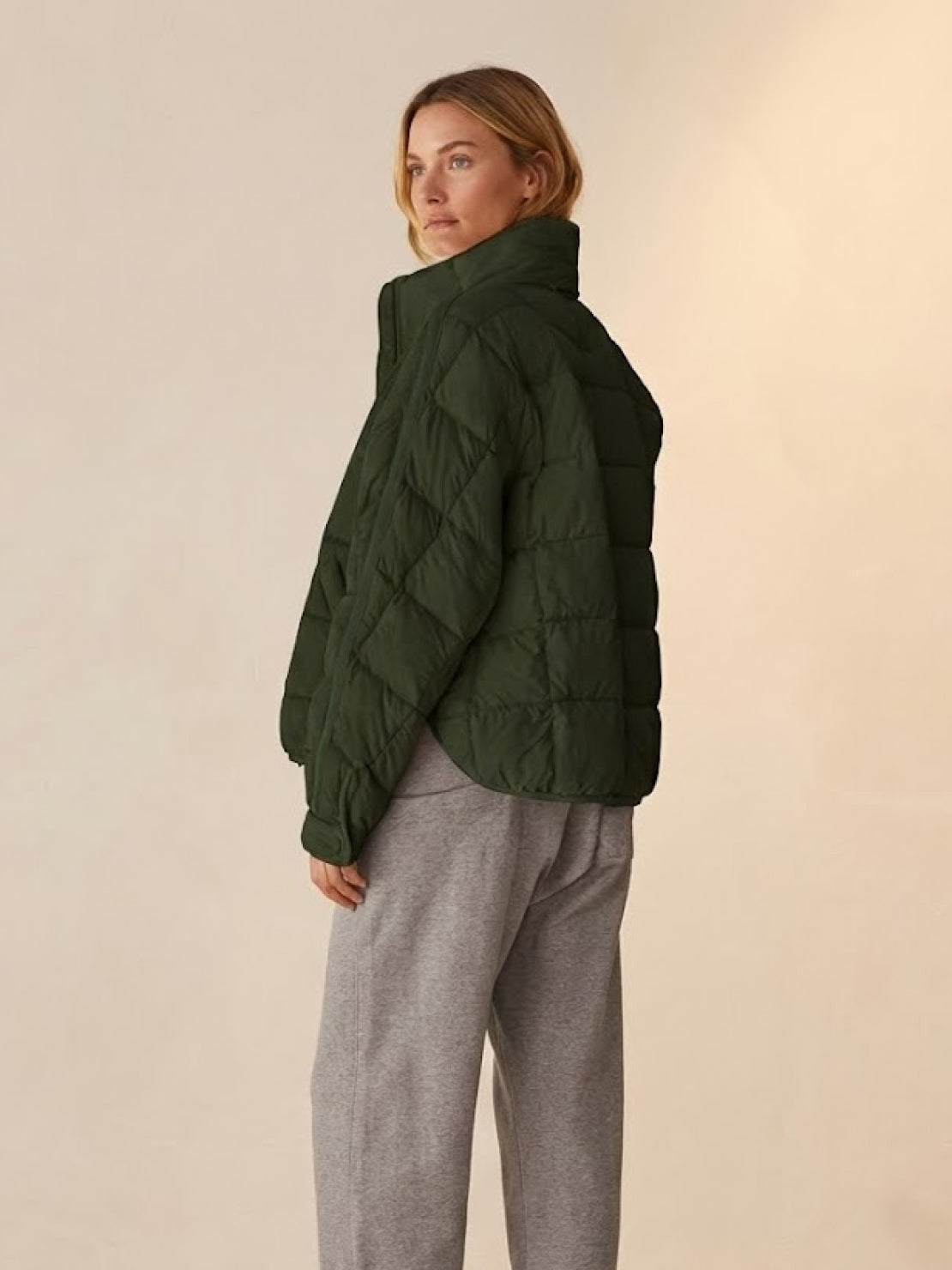 Woman wearing olive green lightweight puffer travel jacket with quilted design and high collar, paired with gray pants.