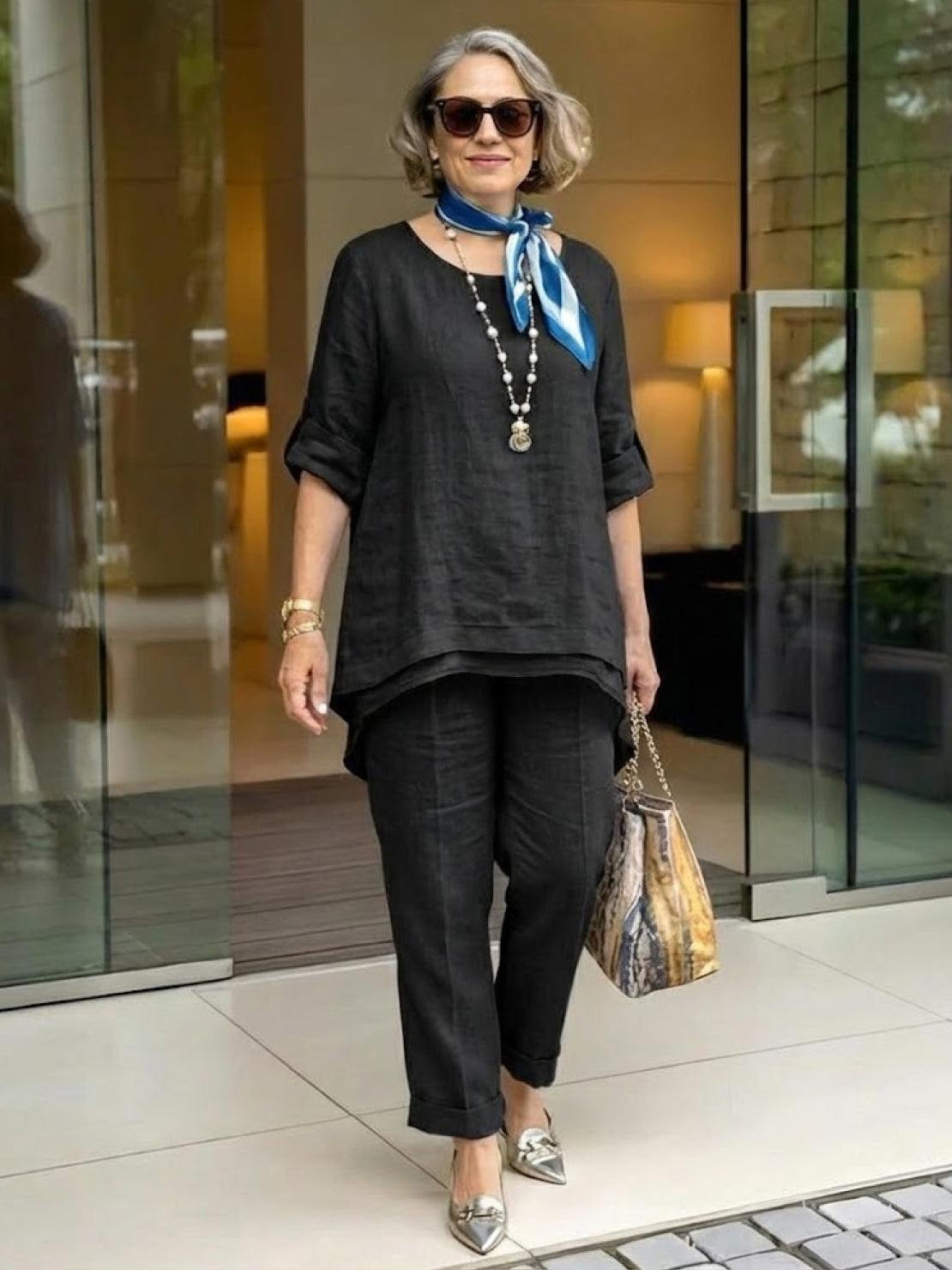 Woman wearing black asymmetrical two-piece trouser set with rolled sleeves and tapered pants, accessorized with scarf and sunglasses.