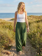 Woman wearing green high-waist palazzo pants and white tank top walking on beach path with grass and ocean background