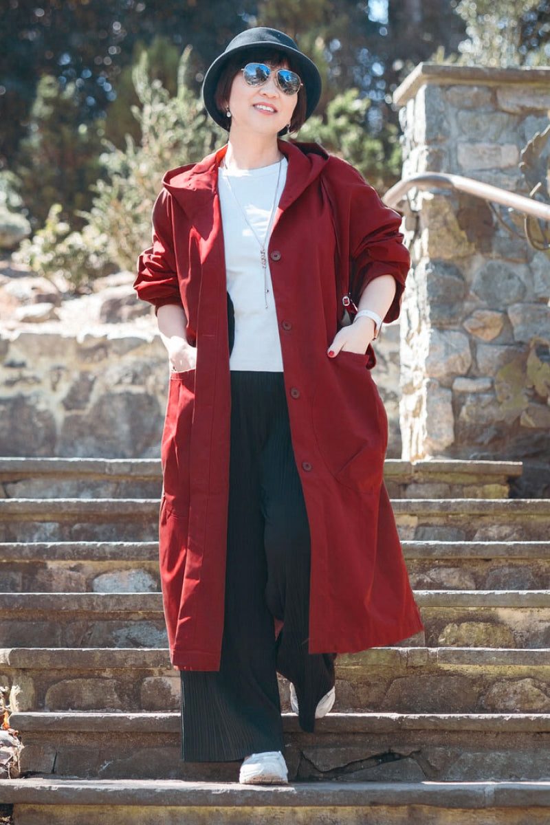 Woman wearing oversized red Heritage Windbreaker Jacket with hood, sunglasses, black hat, white shirt, and black pants outdoors on stone stairs