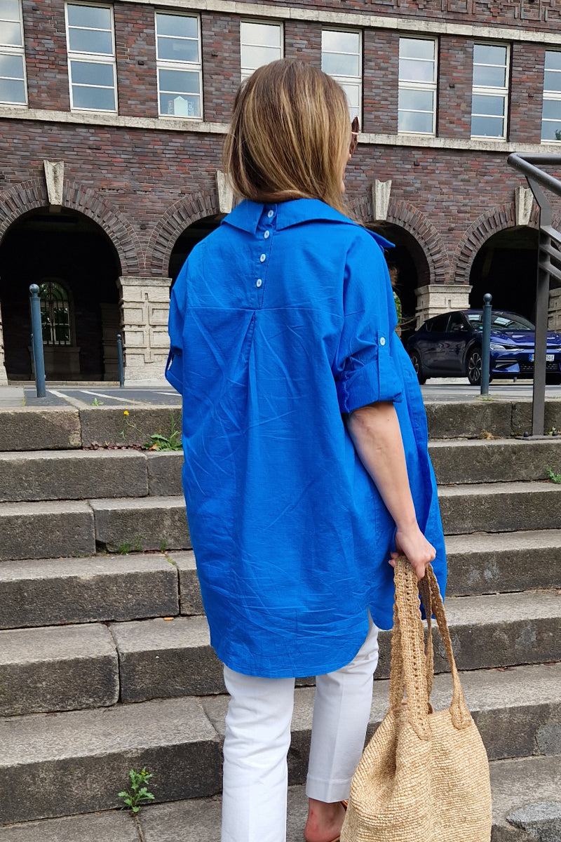 Woman wearing a bright blue oversized linen shirt with back button detailing and rolled sleeves, paired with white pants and carrying a woven bag, standing on outdoor stone steps.