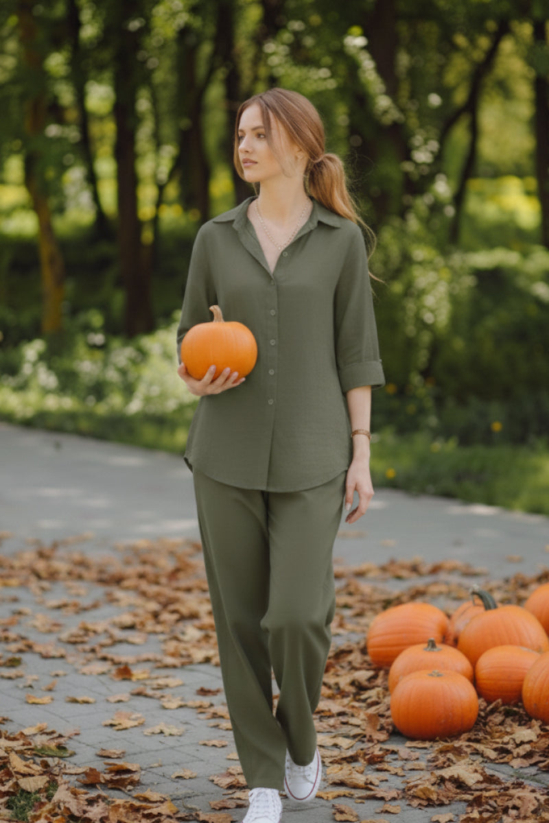 Woman wearing oversized 100% cotton olive green shirt and wide-leg pants holding a pumpkin outdoors with more pumpkins on the ground.