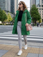 Woman wearing green oversized Rain Couture Windbreaker with hood, black top, gray pants, and white sneakers walking in city crosswalk