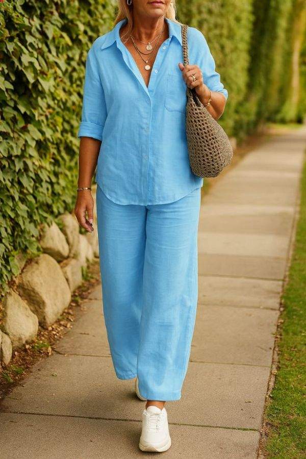 Woman wearing light blue relaxed shirt and trousers two-piece set with rolled-up sleeves and white sneakers walking outdoors